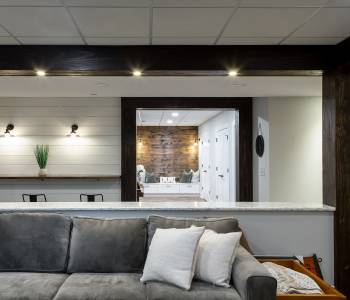 Remodeled basement with white shiplap wall, wood beam across ceiling, and gray couch.