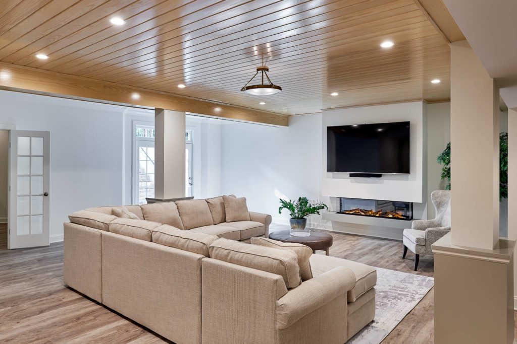 Remodeled basement with wood flooring, off-white sectional couch in the middle, facing mounted TV on far wall. Wood beam ceiling with recessed lighting.