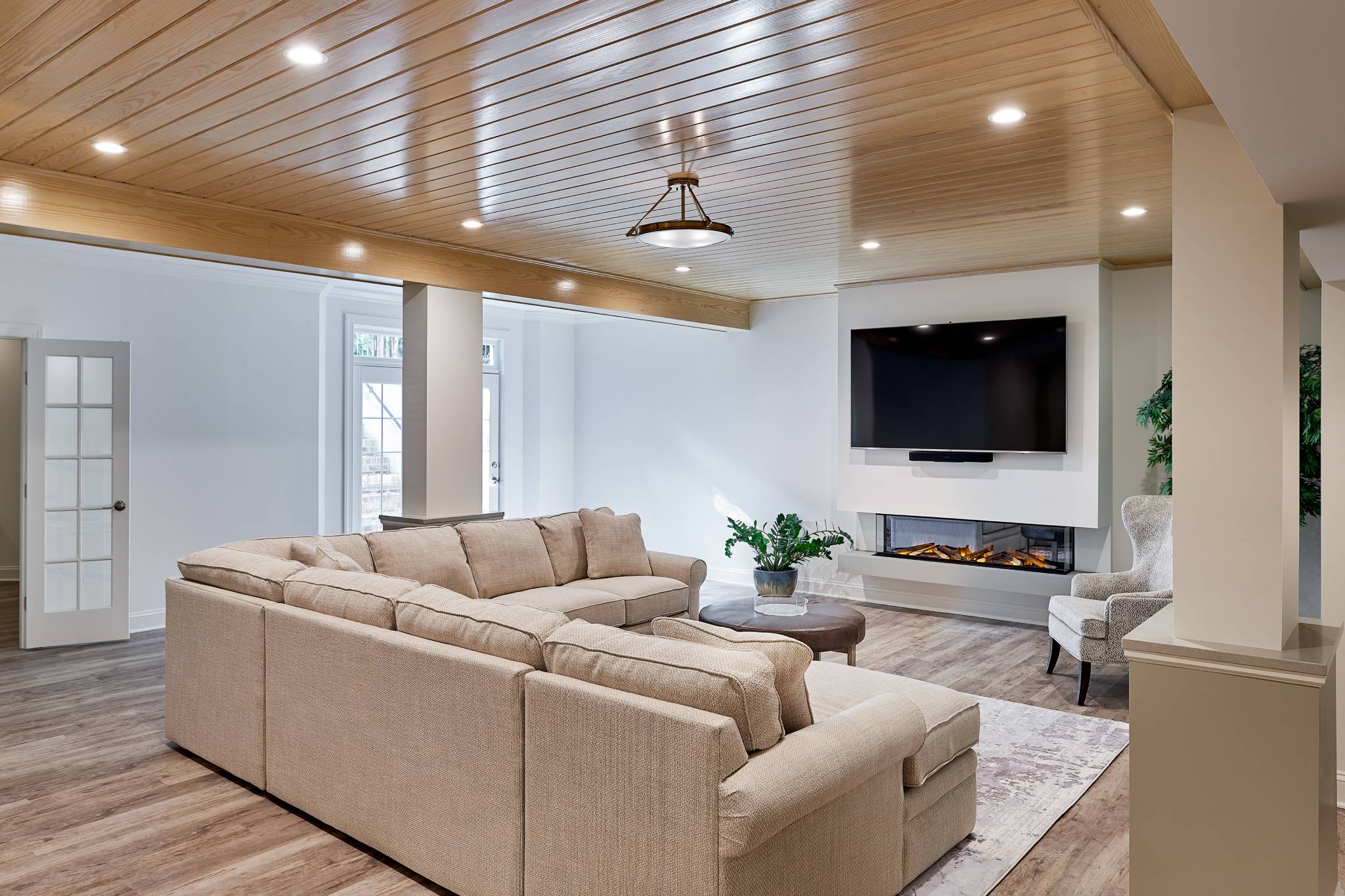 Remodeled living area with light wood floors, woodbeam ceiling with recessed lighting, large tan sectional sofa in middle, facing mounted TV on far wall. White walls. French doors on far wall. Electric fireplace below TV.