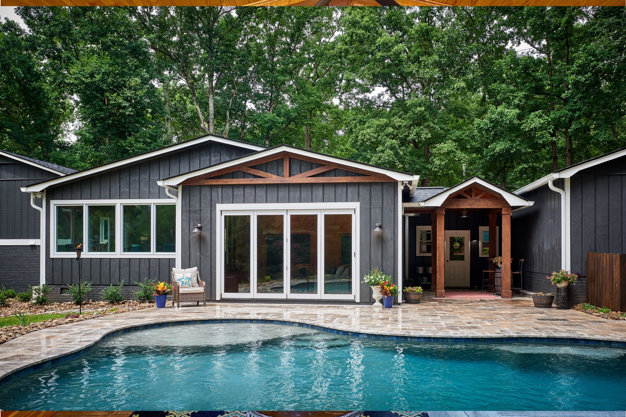 exterior view of blue home with accordion doors leading to large pool and patio in middle of trees
