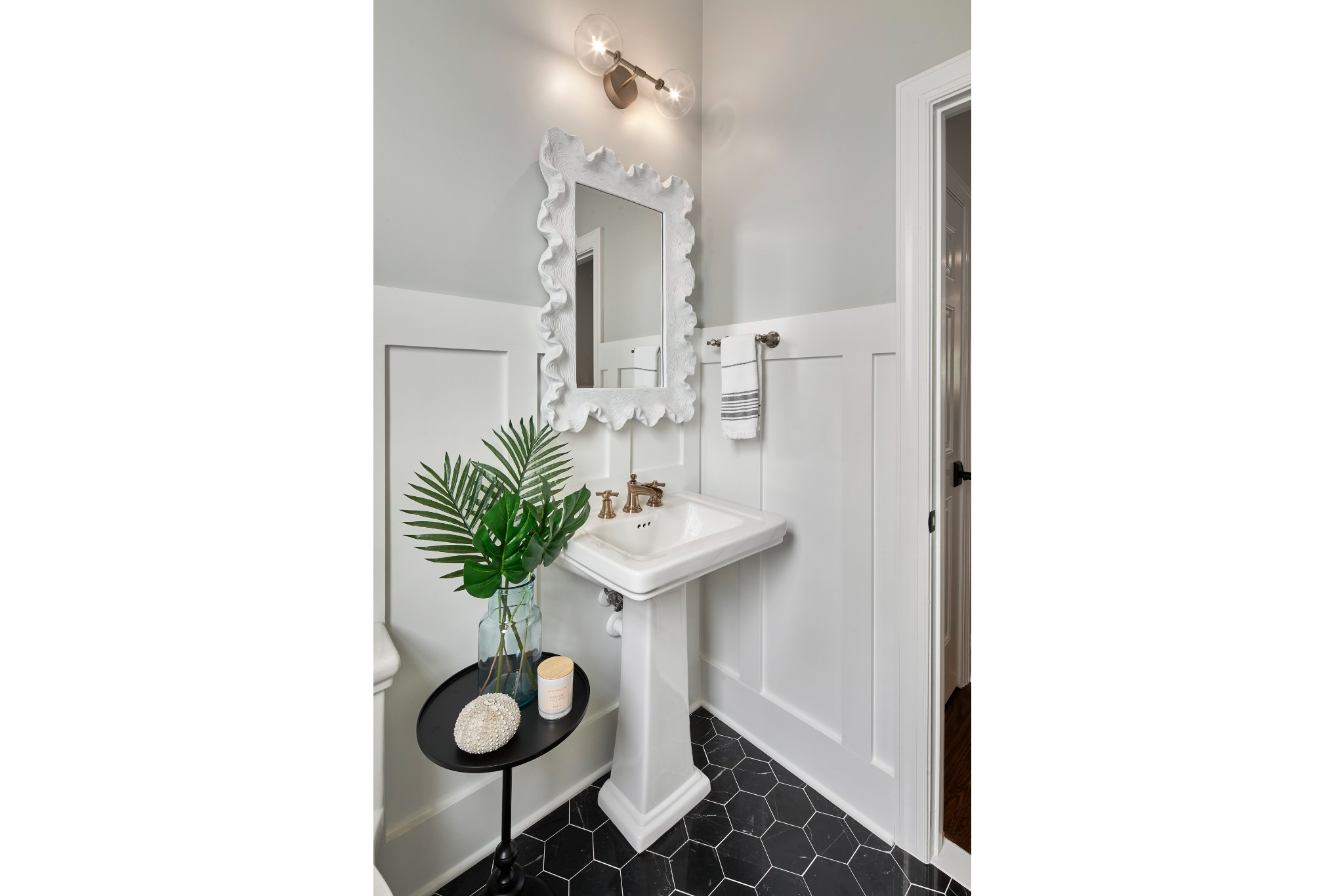 Powder room with white wainscoting and pedestal sink, intricate hanging mirror, black hexagonal tiled floor, and palm frond vase
