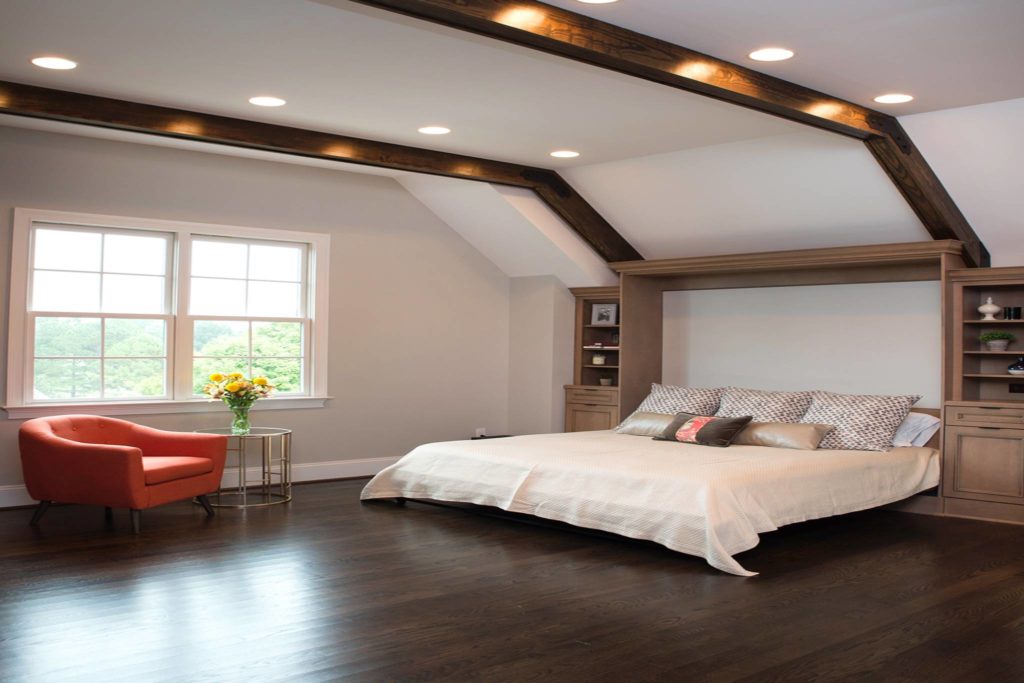 Remodeled primary bedroom with dark wood floors, bed and built-in woodgrain shelving on either side on far right wall, wood beams on ceiling, recessed lighting, two double-hung windows with grids on left wall. Lots of open space.