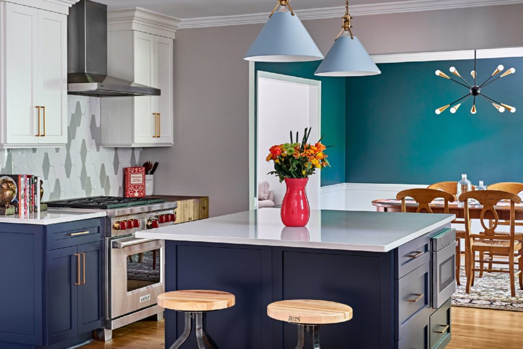 Remodeled kitchen with dark blue lower cabinets, white upper cabinets, white and gray tile backsplash, small square island with stool seating, white stone countertops, open to dining area in background with bright teal wall. Wood floors. Bright pink vase of flowers in middle of kitchen island. Pale blue pendant lights over island.