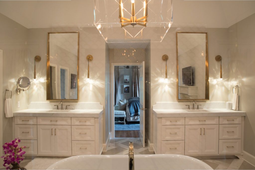 Remodeled bathroom with white dual vanity on either side of open door leading to bedroom. Gold fixtures, stand-alone white soaking tub in foreground.