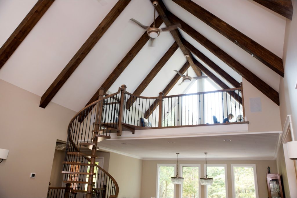 home remodel with exposed ceiling beams over winding staircase and woman working upstairs in loft area.