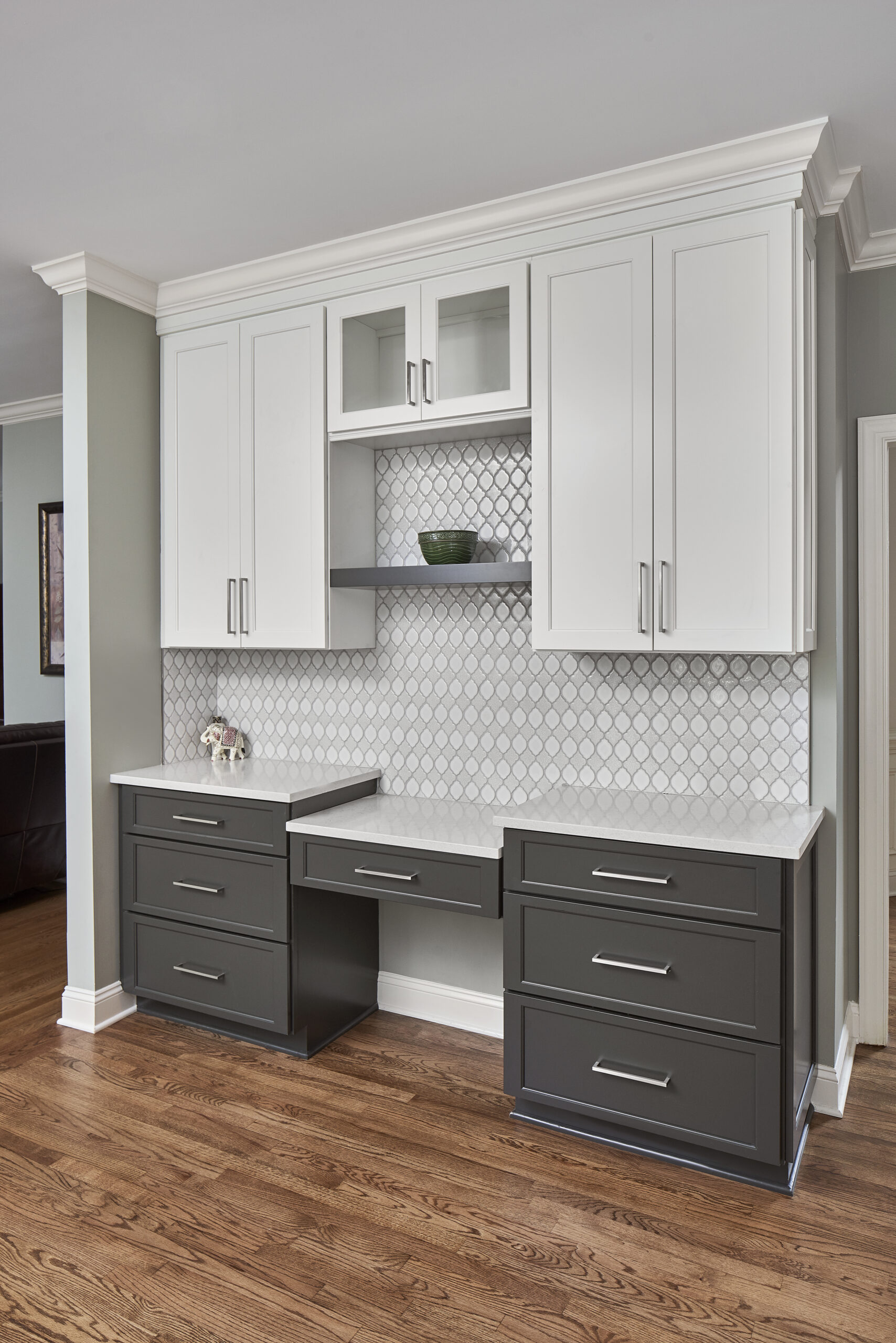 Home office nook with built-in cabinetry, gray lower and white upper, with wood floor, white tile backsplash, and crown molding against ceiling.