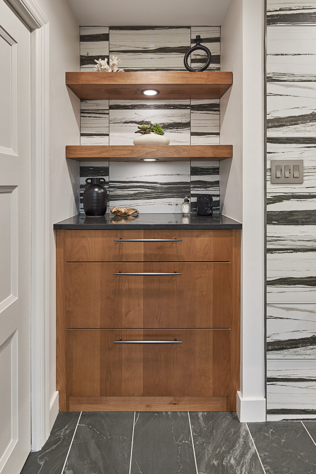 A small nook in a wall if a home, with built-in lower woodgrain cabinet with three drawers and two open shelves above, with accent wallpaper behind them. Dark gray large-tile flooring.