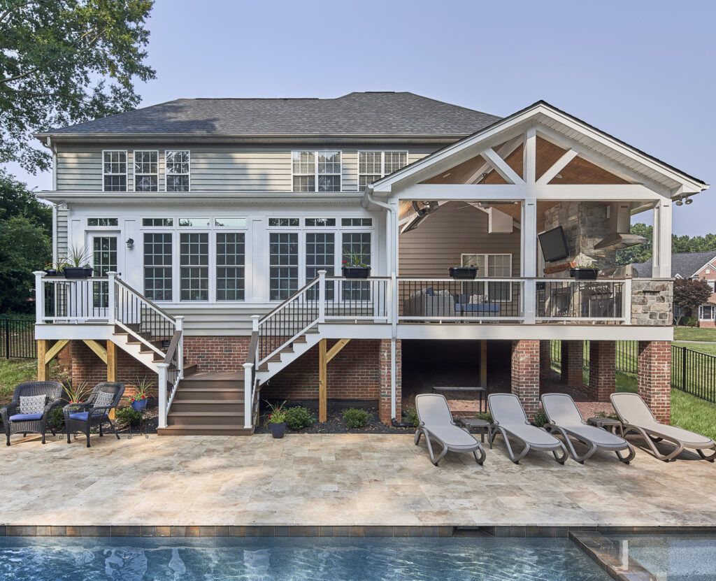 Back of a remodeled home with covered upper deck with stairs leading down to patio area around an inground pool.