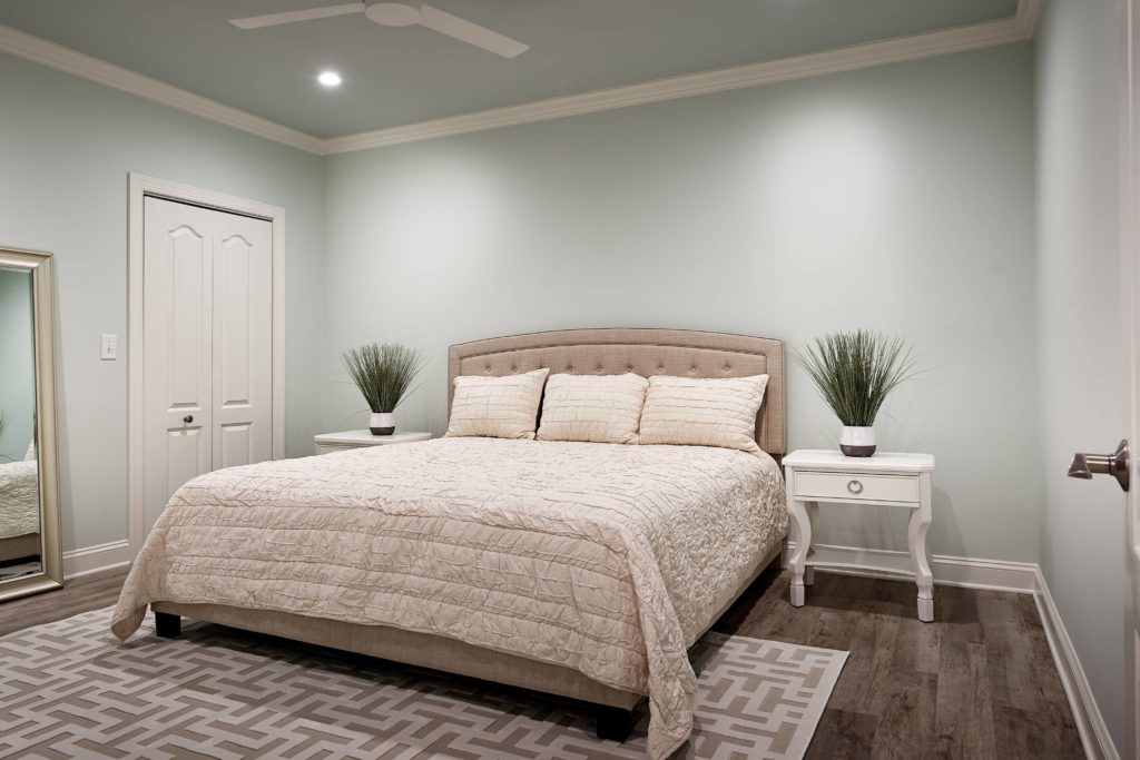 Primary bedroom remodel with gray wood flooring, queen bed with white comforter in the middle, with small white nightstand to the right. White closet door on the left wall. Pale green/blue walls with white trim and small white ceiling fan.