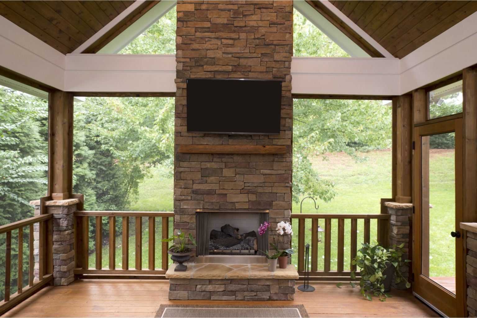 Three-season room with large windows, stone fireplace, and TV. Looking out on wooded backyard.