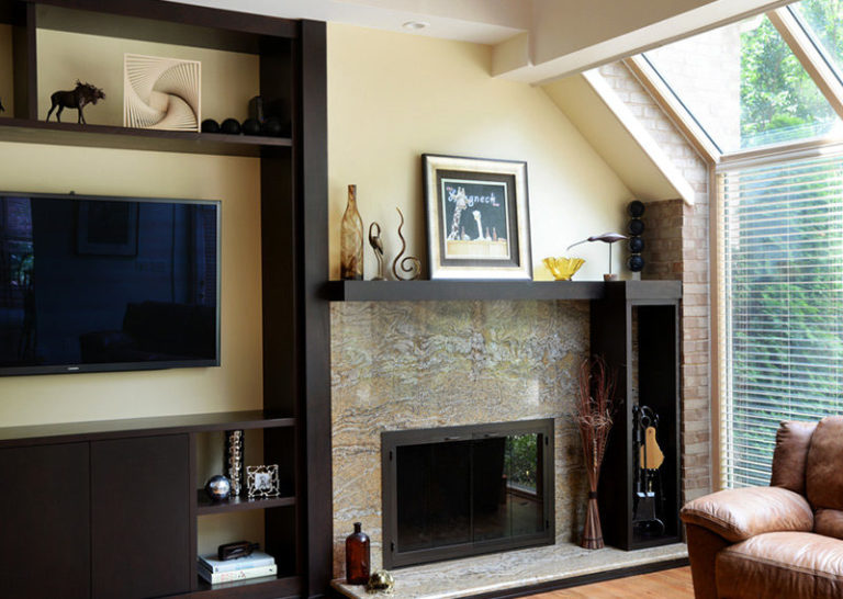 A modern gas fireplace with stone surround and black mantel shelving against pale yellow wall in living area with floor-to-ceiling window to the right with view of green trees.