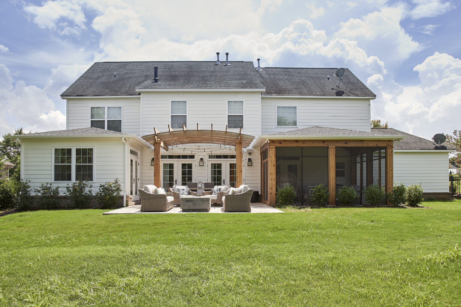 Screened-in porch addition on back of large two-story white home, leading out to patio area with wooden pergola and cushioned patio furniture. Large mowed green lawn in foreground. Blue sky and clouds behind house.