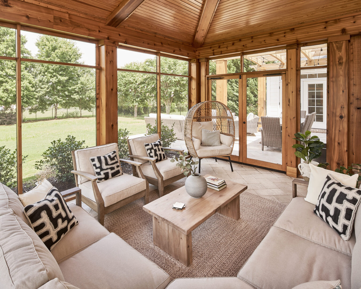 Interior of a screened-in porch with woodgrain vaulted ceiling, light gray/tan tile floor, white and light wood patio furniture, wood-paneled walls, double glass doors leading to patio on far wall.