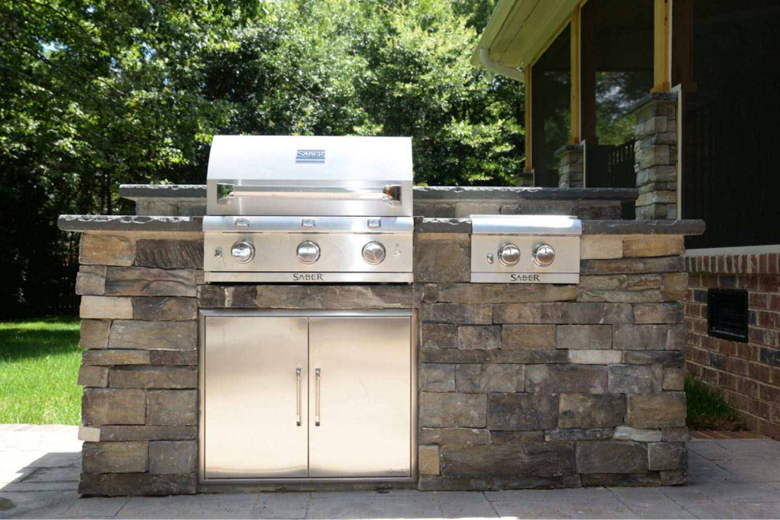 Modern stainless steel grill built into stone countertop outside a brick home.