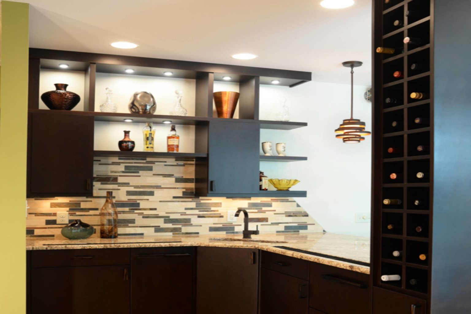 Modern kitchen with black cabinets and tall wine-bottle shelving on the right, with in-shelf lighting.
