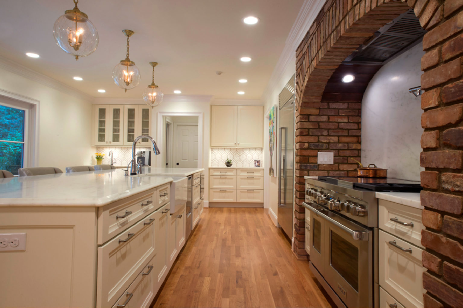 Remodeled white kitchen with large island and gold pendant lighting. Wood floors. Arched brick around the cooktop area.