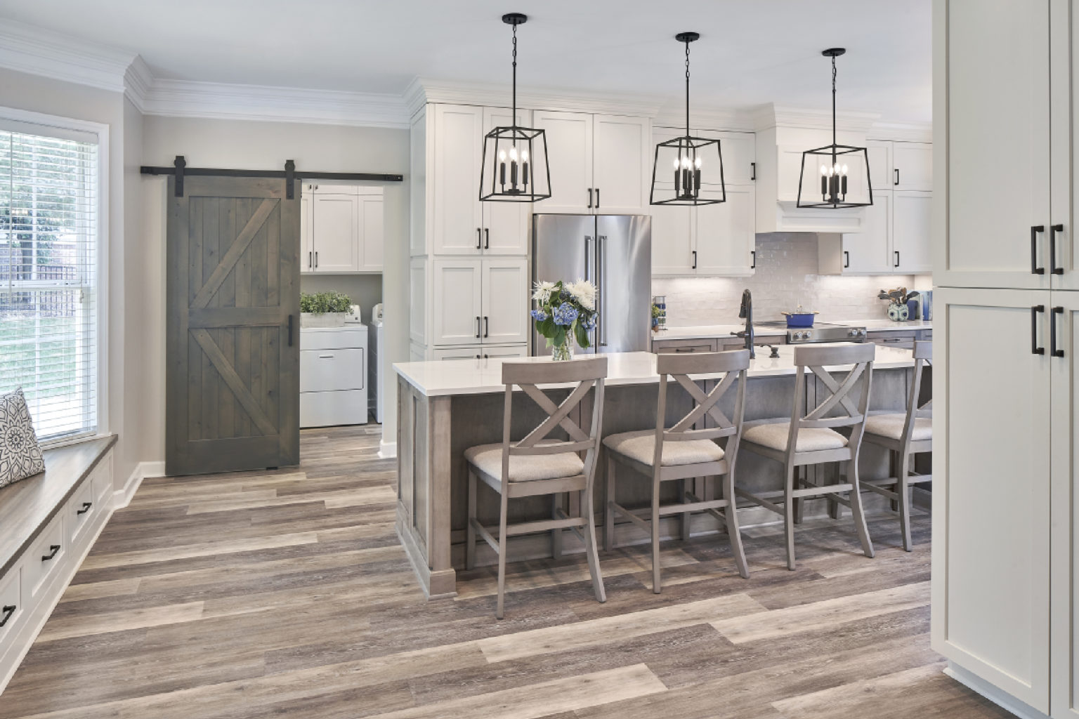 Remodeled gray-and-white kitchen with stools at the island, black pendant lights, and gray barn door in background, open to laundry area.