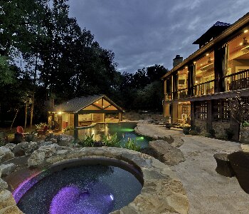 Luxurious backyard remodel with stone ground, in-ground pool, and pool house addition, at dusk. Lights on inside house and pool house.