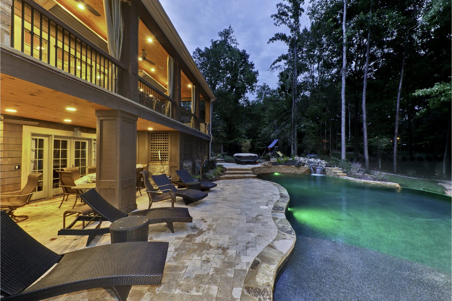 Back of a large home at night, with in-ground pool on the right and two-level patio/deck on back of the house, on the left.