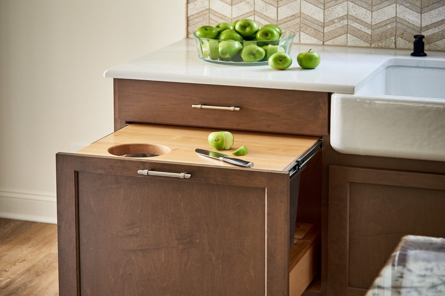 Wood pull-out cutting board with a peeled green apple and pearing knife on it. Woodgrain cabinets, white stone countertops.