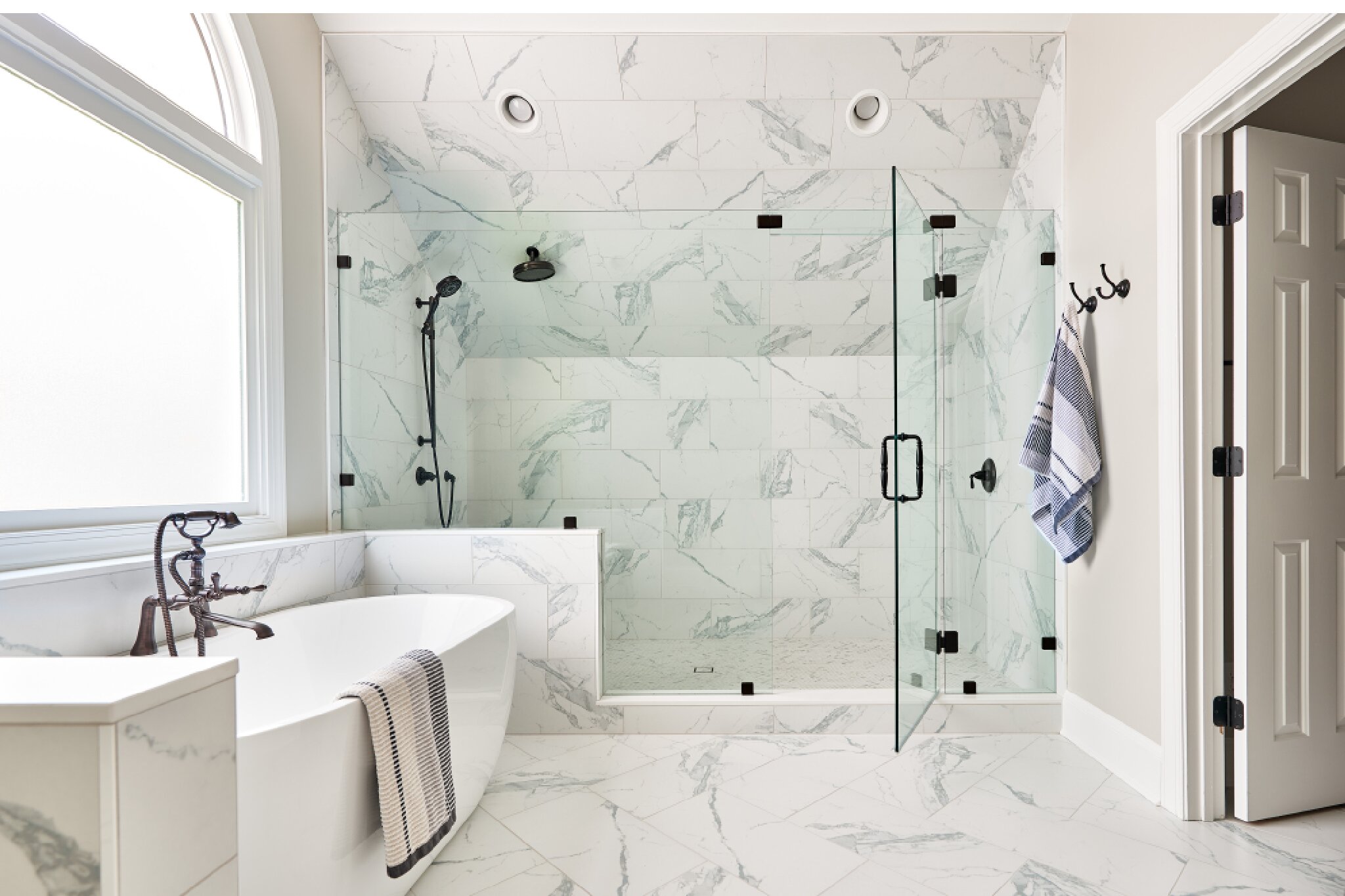Bathroom remodel with white marble floors, glass-enclosed shower on far wall with black fixtures, white soaking tub on left wall, under large picture window.