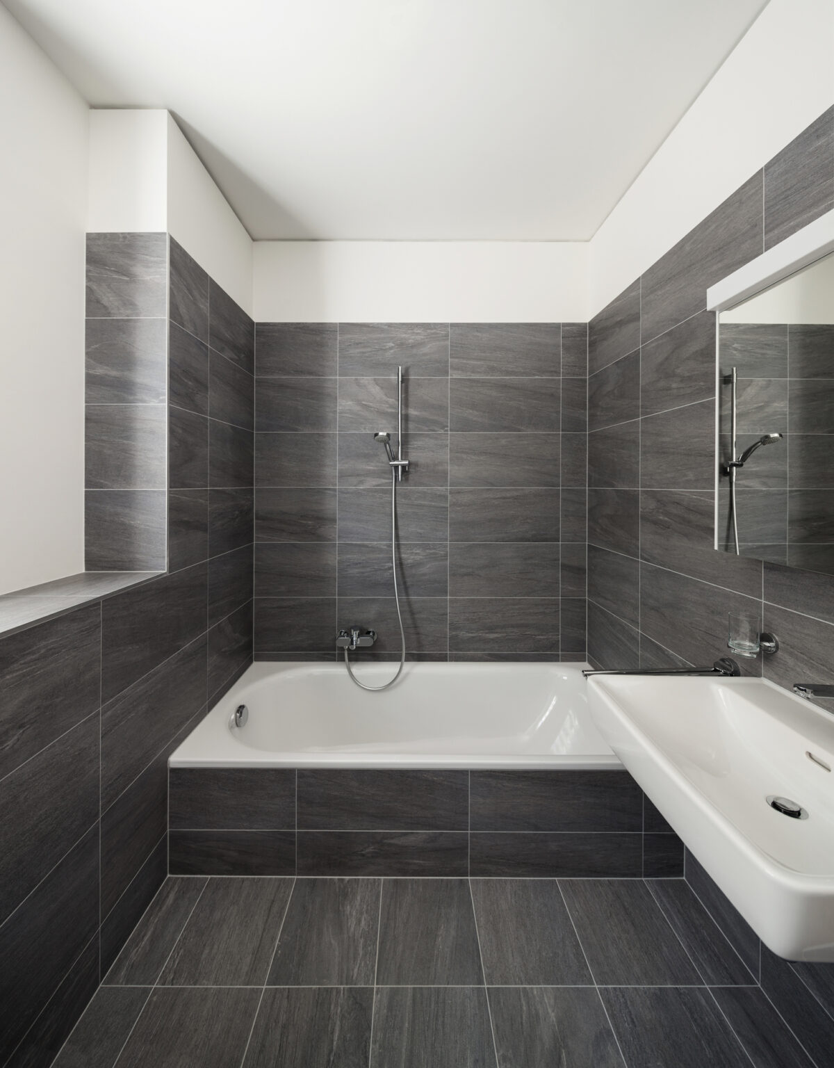 Interior of a modern house, gray bathroom, tiled walls and white ceiling, and alcove tub on far wall.