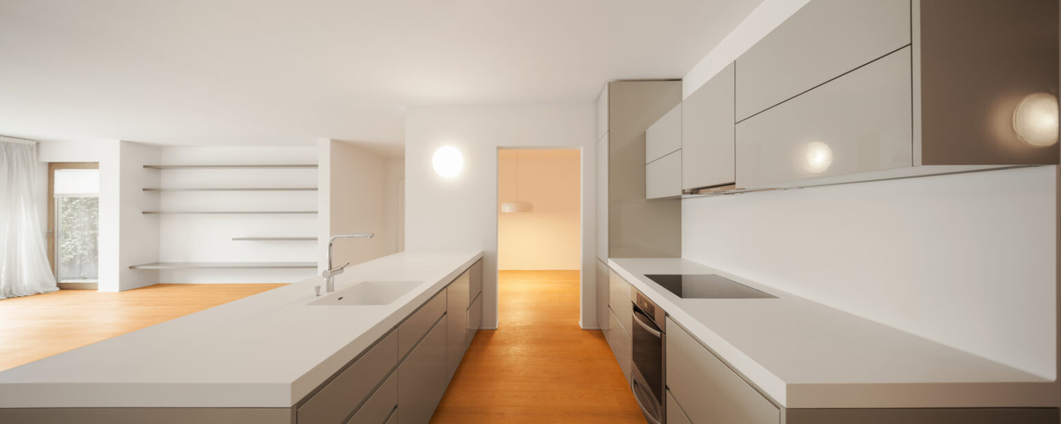 Galley kitchen with light wood floors, white/gray modern cabinets, white stone countertops, open to empty living area on the left.