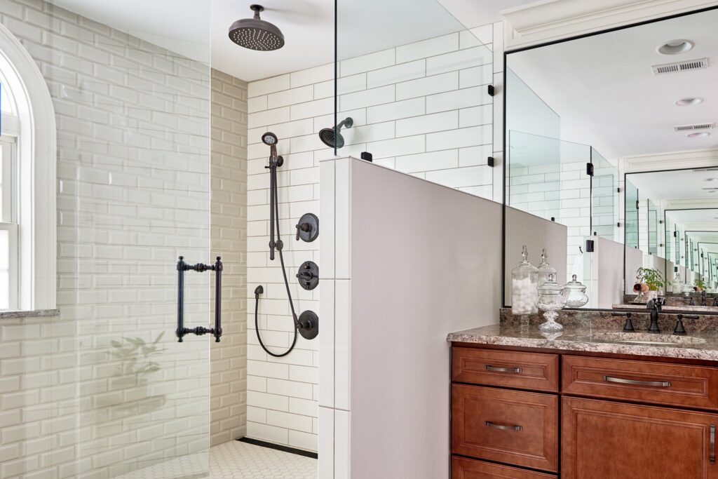 Remodeled bathroom with glass-enclosed shower with white subway tile walls, black fixtures, woodgrain vanity with stone countertop and large mirror on wall.