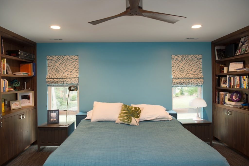 Renovated primary suite with sky blue accent wall, bed with blue comforter in the middle, built-in dark wood shelving on either side, and small ceiling fan.
