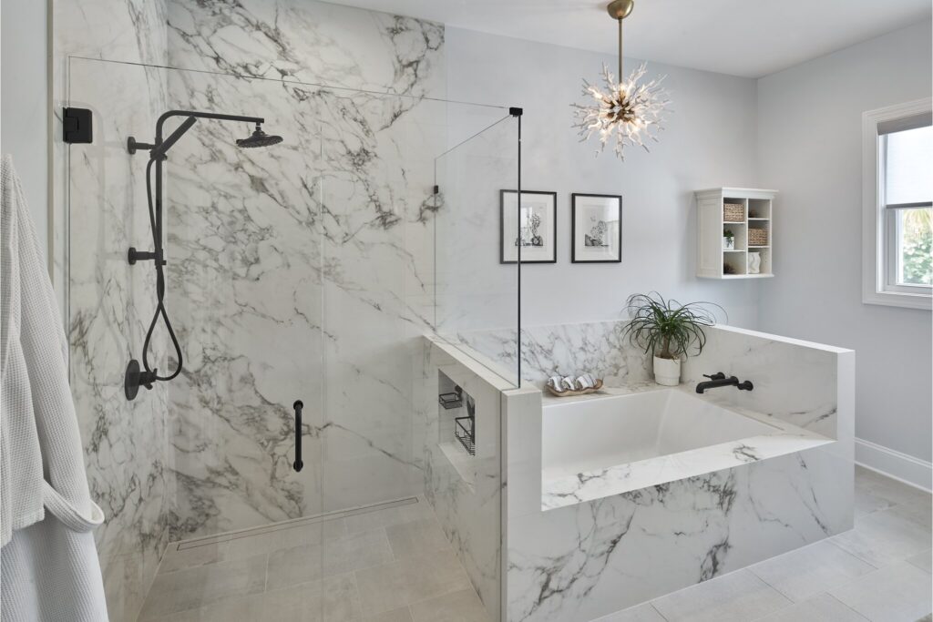 Modern bathroom remodel with white marble shower walls and surrounding stand-alone tub. Black showerhead and fixtures. White walls and white tile floor.