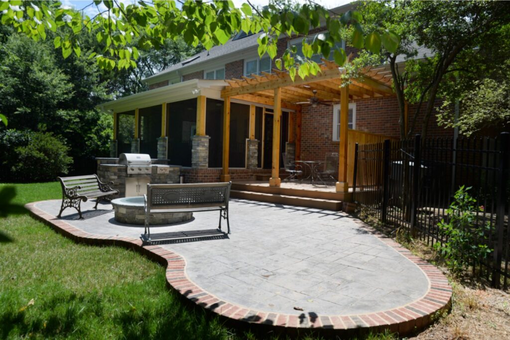 Exterior remodel featuring stone paver patio in unique rounded natural shape, with built-in stone firepit on the far end, a large stainless steel grill, and two benches on either side of the fire pit. Large brick home with screened-in porch and pergola behind the patio. Trees surrounding.; Outdoor patio with a grill and ample seating around a built-in round stone firepit, behind a large two-story brick home, with trees surrounding.