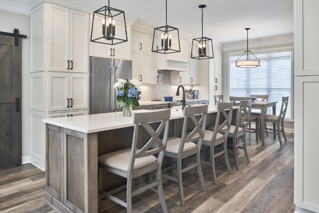 Remodeled kitchen with long island with gray stool seating, white stone countertop, white cabinets along wall behind. Gray/wood flooring, pendant lights.; post img