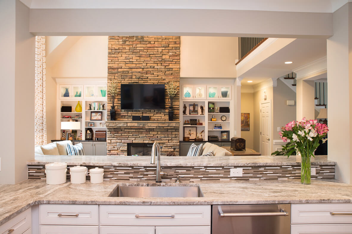 Remodeled kitchen with white cabinets and light stone countertops, open to living area beyond, with tan brick fireplace and white built-in shelving on either side.